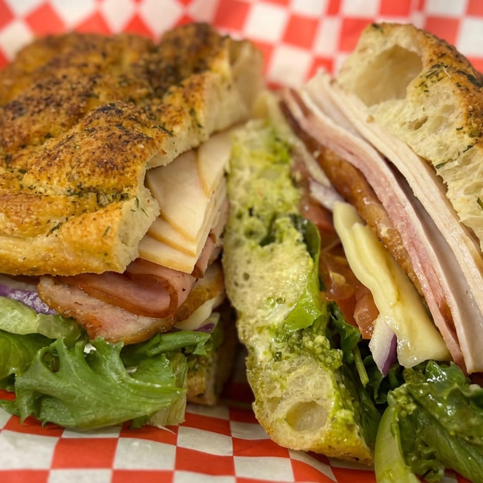 Loaded focaccia sandwich with sliced deli meats, cheese, lettuce, tomato, onion, and avocado on red-and-white checkered paper.