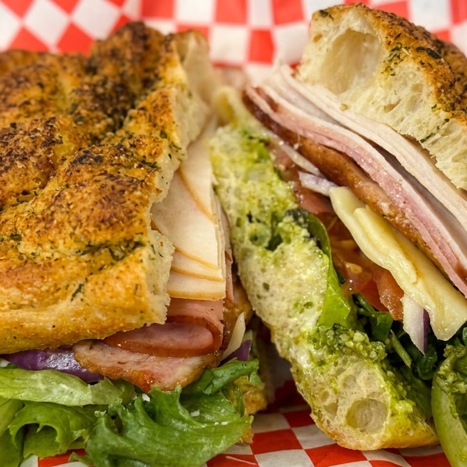 Close-up of a toasted Italian sandwich with ham, cheese, lettuce, onions, and peppers on red-and-white checkered paper.
